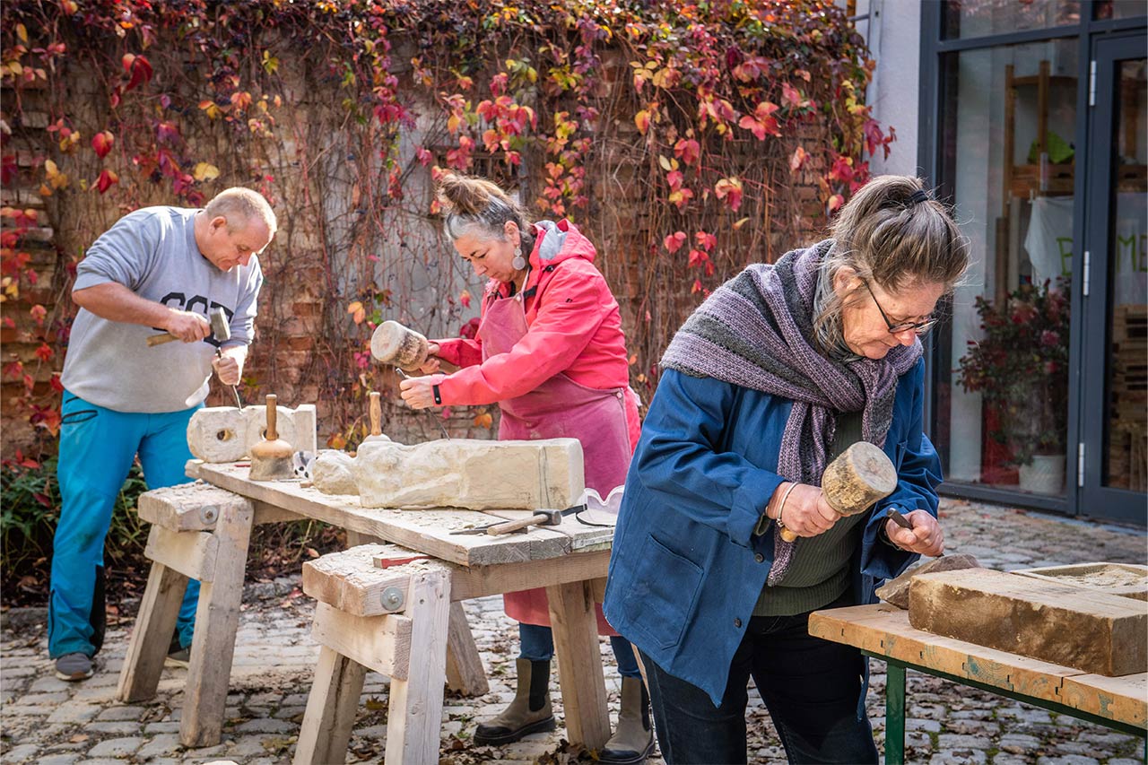 Drei Personen arbeiten konzentriert im Freien an Werkbänken aus Holz. Sie bearbeiten helle Steinblöcke mit Hammer und Meißel. Im Hintergrund ist eine mit roten und grünen Weinblättern bewachsene Mauer zu sehen. Die Szene zeigt eine kreative Arbeitsatmosphäre beim Seminar „Figürliches Gestalten in Stein“ der Kulturakademie Naumburg.